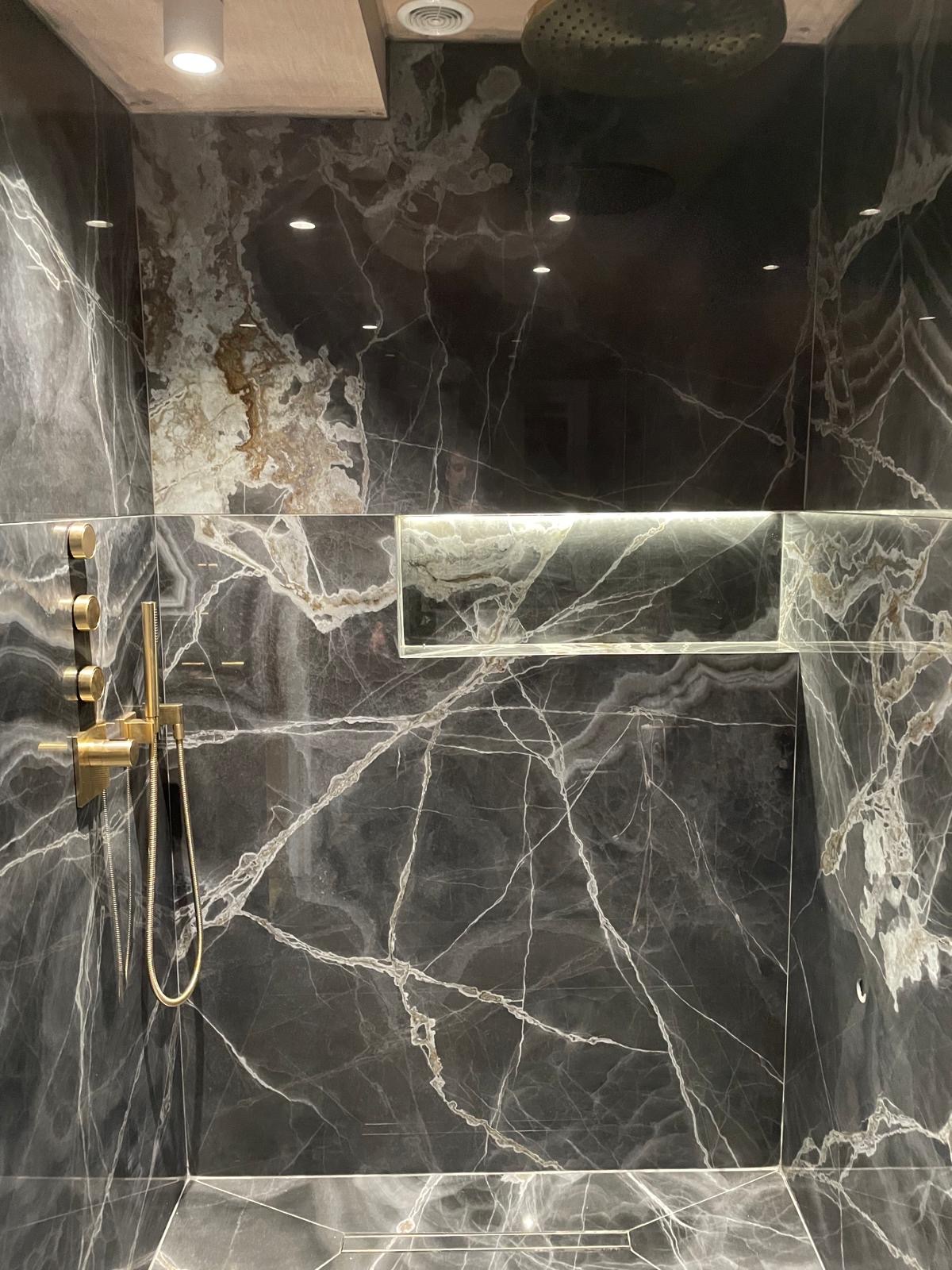 Walk-in shower clad in large-format black marble-effect porcelain with white veining, an LED-lit niche and brushed brass fittings