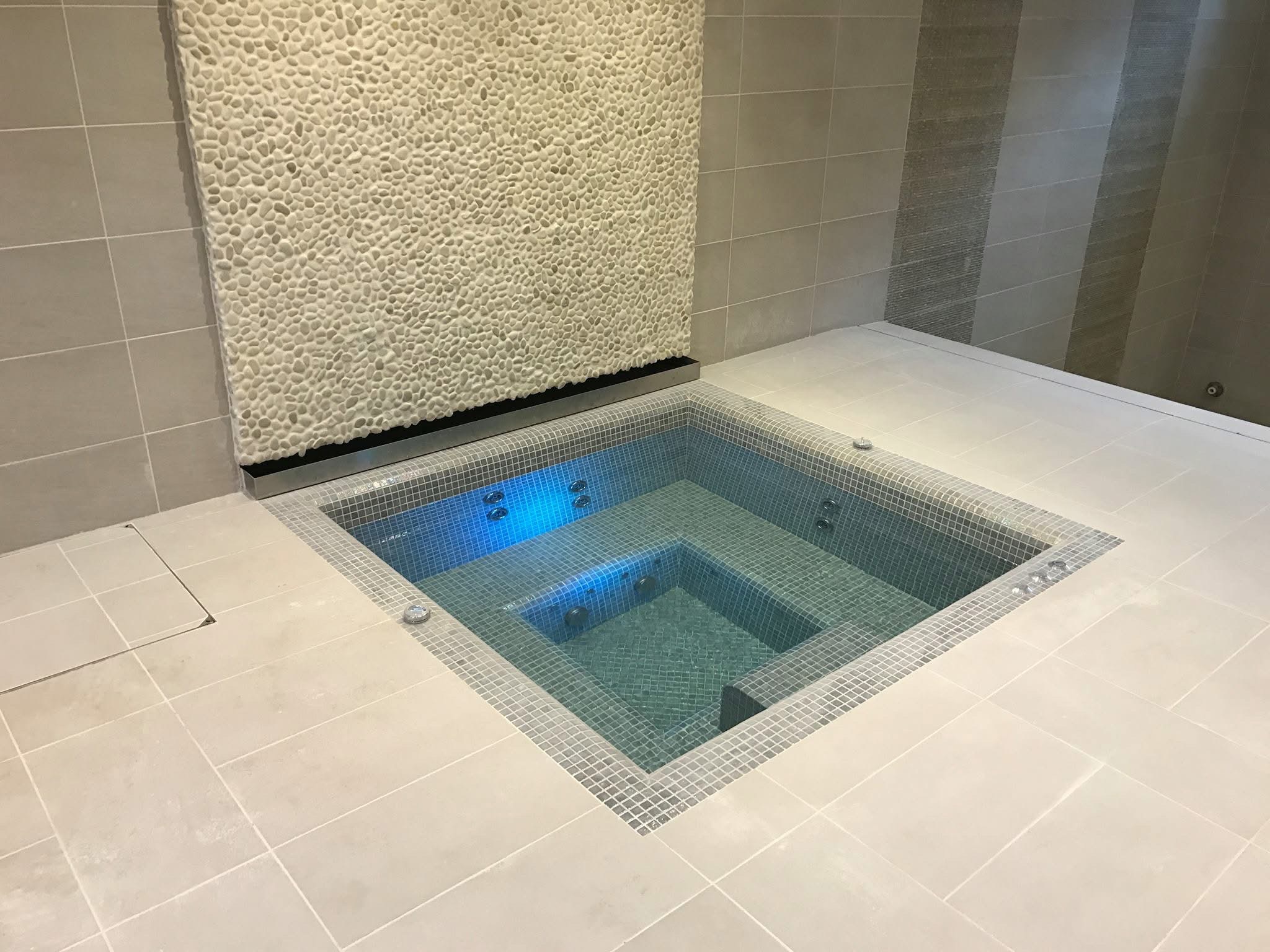 Sunken jacuzzi finished in grey glass mosaic beside a white pebble feature wall and pale porcelain floor